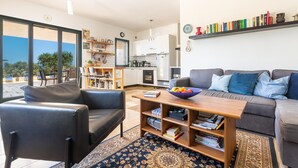A modern living room with a sofa, a coffee table, and a bookshelf. There is a kitchen in the background with white cabinets and a refrigerator. - 2968 Villa Freude by Perle di Puglia (Carovigno)