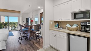 A modern kitchen with white cabinets, a microwave, and a coffee maker. There is a dining area with wicker chairs and a table. - Studio @ Luau! Walk to beach! Sleeps 4~ (Miramar Beach)