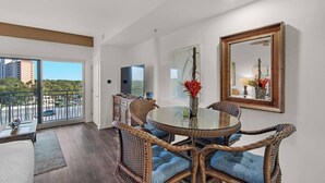 A modern living room with a glass dining table surrounded by wicker chairs, a flat-screen TV, and a large mirror on the wall. - Studio @ Luau! Walk to beach! Sleeps 4~ (Miramar Beach)