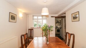 Un coin repas avec une table en bois, des chaises, un vase de fleurs, une fenêtre avec des stores et, au fond, une cuisine.