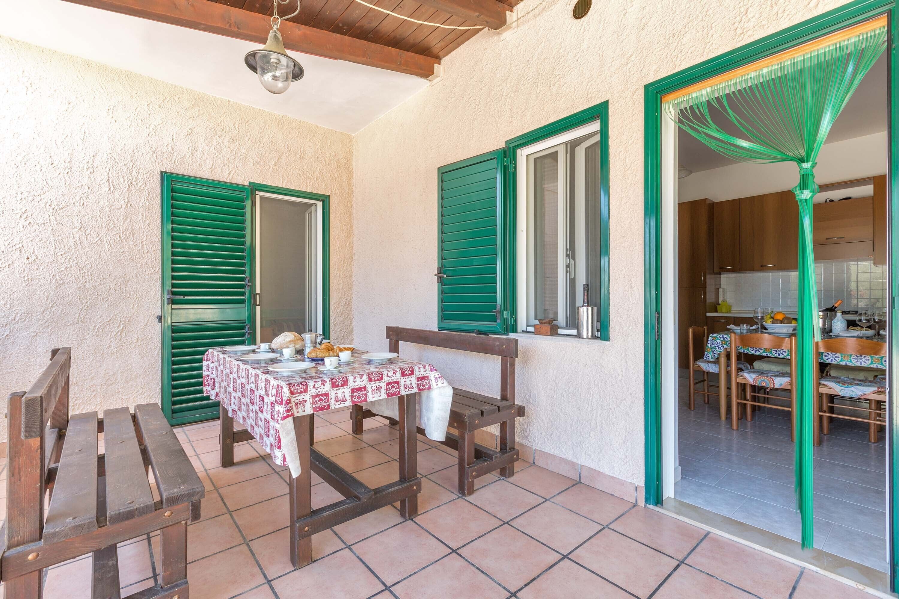 Une terrasse avec une table et des chaises en bois, une porte verte et une vue sur un coin cuisine.