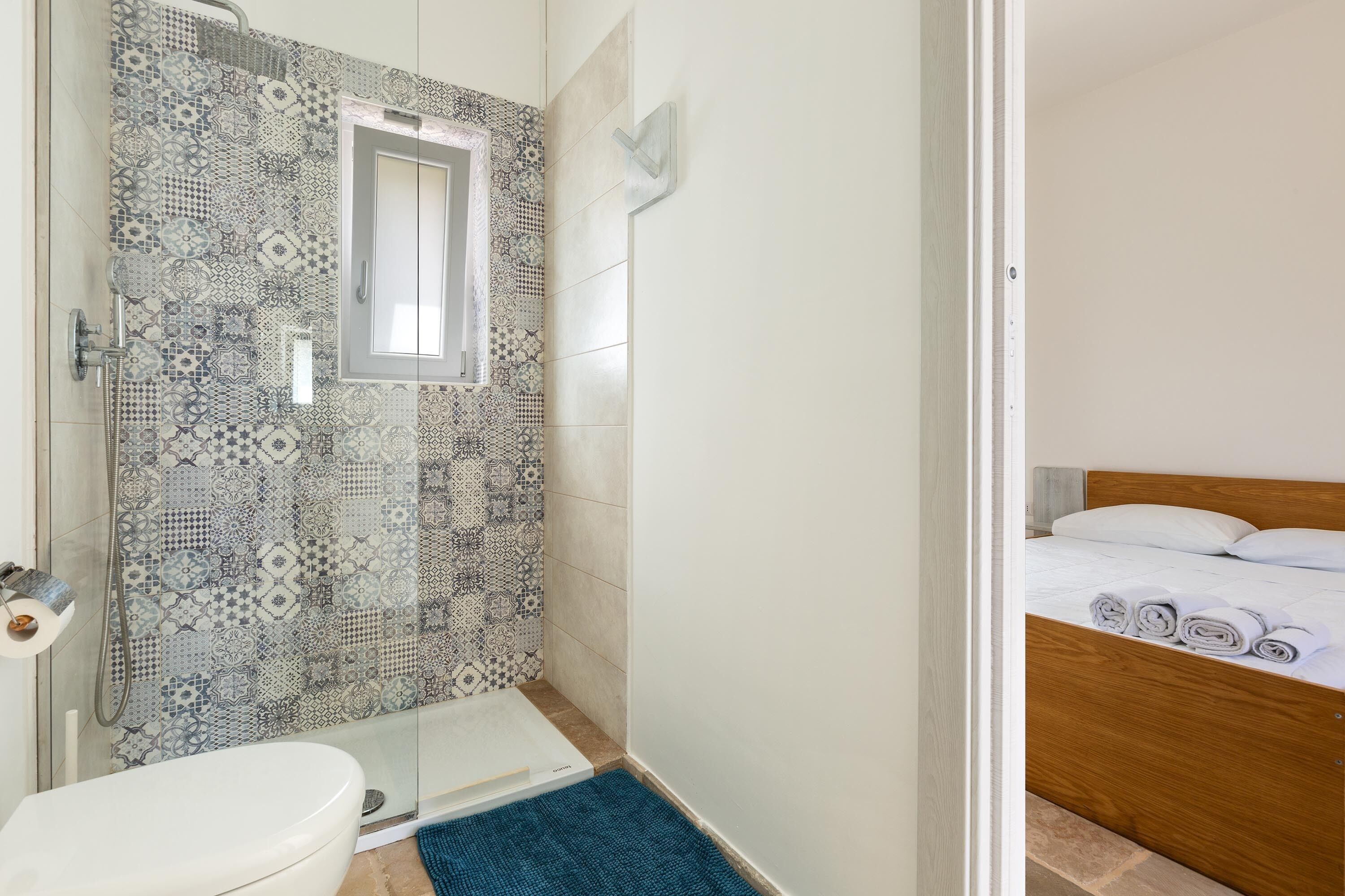 A bathroom with patterned tiles, a shower, a toilet, and a bed with rolled towels.
