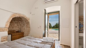 A bedroom with a stone fireplace, a bed, a wooden dresser, and a view of a pool through an open door. - 2329 Villa Kairos by Perle di Puglia (Ostuni)