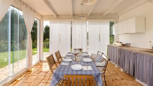 A dining area with a table set for four, surrounded by chairs, a kitchen area with cabinets, and large windows offering a view of the outdoors. - 2329 Villa Kairos by Perle di Puglia (Ostuni)
