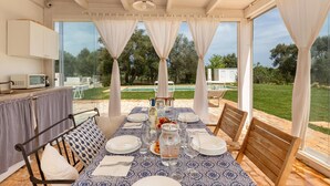 A covered outdoor dining area with a table set for four, a grill, and a view of a pool and garden. - 2329 Villa Kairos by Perle di Puglia (Ostuni)