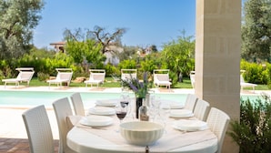 An outdoor dining area with a pool view, white wicker chairs, and a table set for six with wine glasses and a bowl. - 2921 Villa La Seta by Perle di Puglia (San Vito dei Normanni)