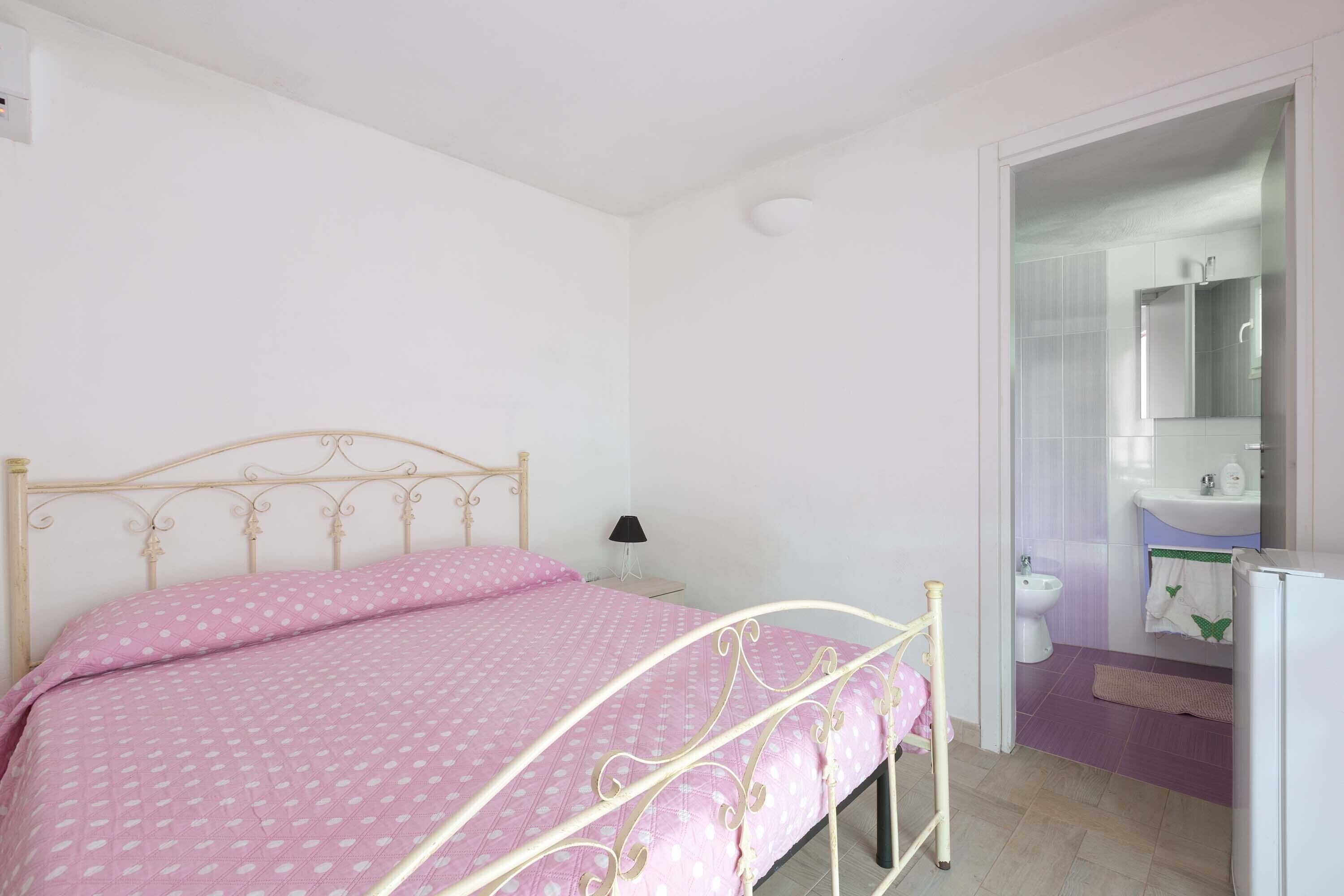 Ein Schlafzimmer mit Bett, Nachttisch, Lampe und einem durchgehenden Türspalte sichtbaren Badezimmer.