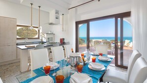 Een eetgedeelte met een tafel voor vier personen, een keuken op de achtergrond en uitzicht op zee door grote ramen.