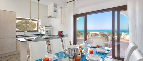 A dining area with a table set for four, a kitchen in the background, and a view of the sea through large windows.