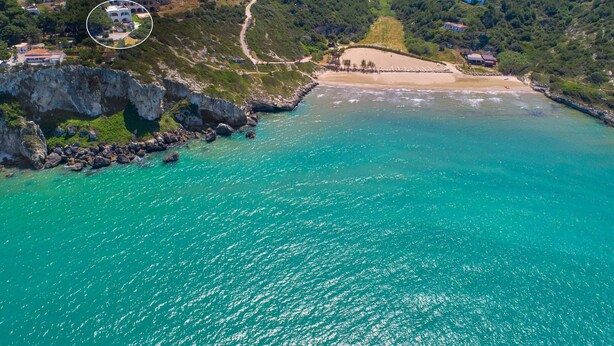 Luchtbeeld van een kustgebied met een strand, rotskliffen en een klein groepje gebouwen.