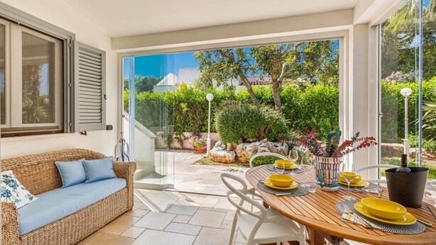 Un patio bañado por el sol con un sofá de mimbre, una mesa de comedor de madera dispuesta con platos y vasos amarillos, y vistas a un exuberante jardín a través de grandes puertas de cristal.