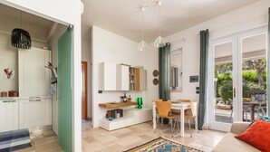 Una sala moderna con un sofá, un comedor con mesa y sillas, una cocina con armarios blancos y un ventanal corredizo que da a un espacio exterior.