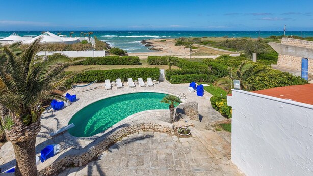 A pool area with clear blue water, surrounded by lounge chairs and palm trees, overlooking a beach and ocean. - 2374 Villa Costa Merlata by Perle di Puglia (Ostuni)
