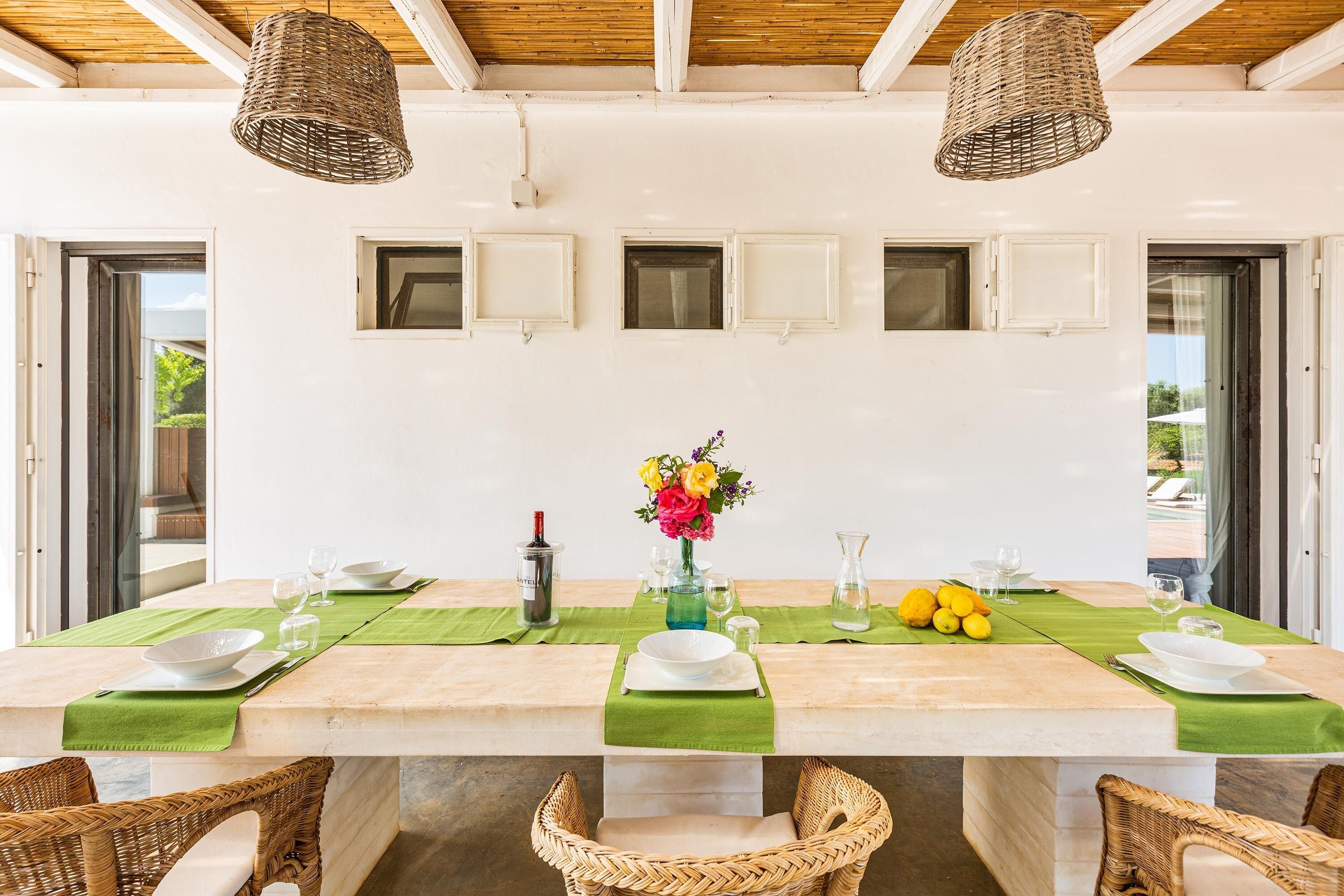 A dining area with a long table set for four, surrounded by wicker chairs, with a green table runner, a vase of flowers, and lemons on the table.