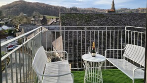 Ein Balkon mit einem weißen Metalltisch und -stühlen, einer Champagnerflasche und einem Blick auf einen Kirchturm in der Ferne.