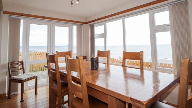 A dining area with a wooden table and chairs, large windows with a view of the sea, and a ceiling light fixture. - Learig (Buckie)