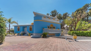 A two-story building with a blue facade, a balcony with yellow railings, and a paved courtyard with potted plants.