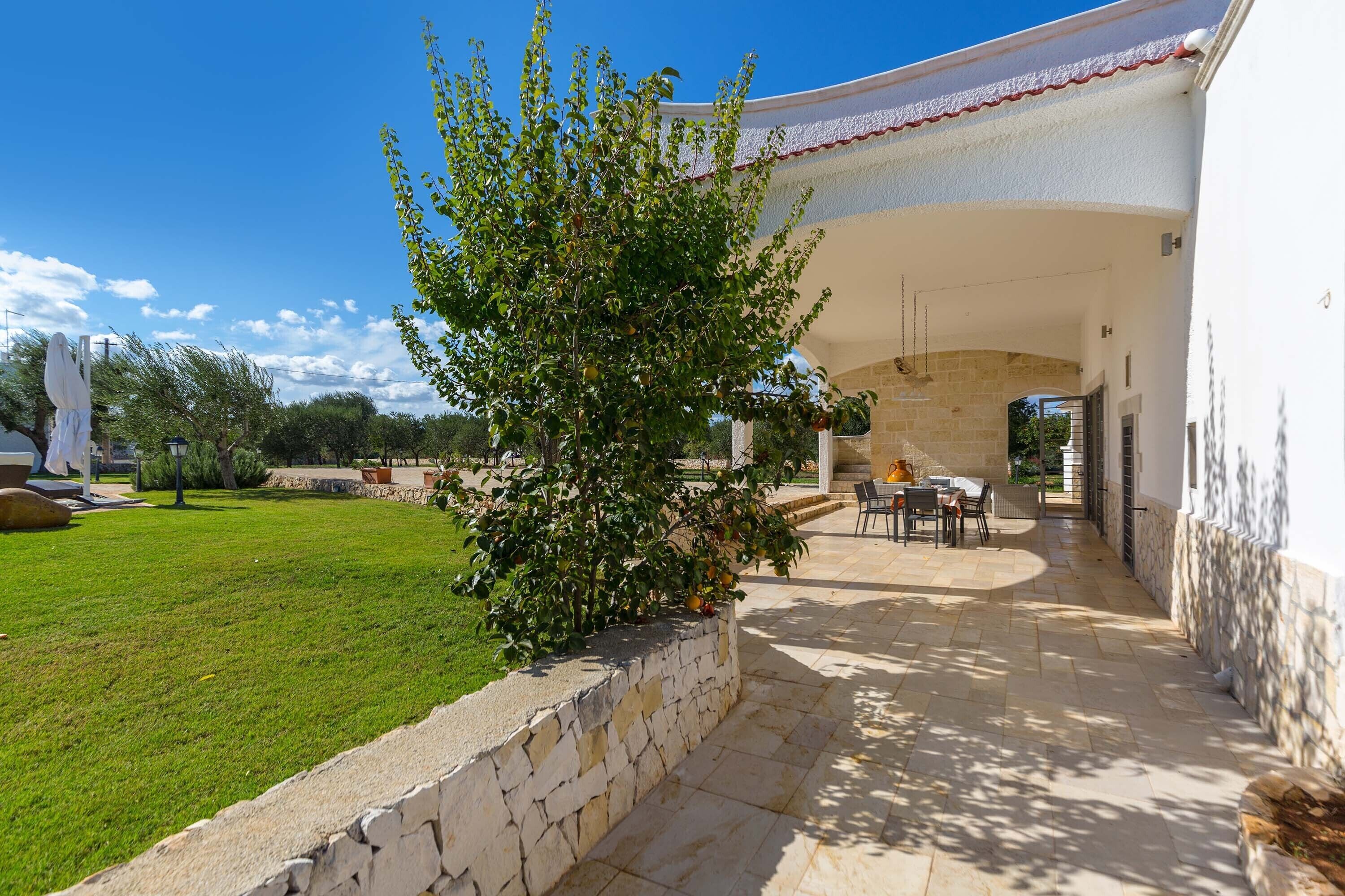 A covered outdoor patio with stone flooring, a stone wall, and a small tree.