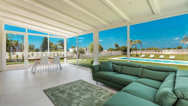 A spacious living room with a green sofa, a dining table with chairs, and large windows offering a view of a pool and greenery. - 3088 Villa Alceste by Perle di Puglia (San Vito dei Normanni)