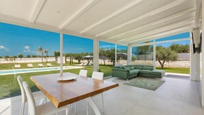 A spacious living area with a wooden dining table, white chairs, a green sofa, and large windows offering a view of a pool and garden. - 3088 Villa Alceste by Perle di Puglia (San Vito dei Normanni)