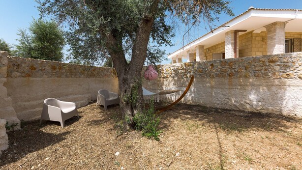 Une cour bordée d’arbres, avec un mur en pierre, un hamac et deux chaises en plastique blanches.
