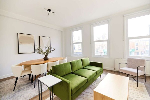 A modern living room with a green sofa, white chairs, a wooden coffee table, and a dining table. There are two large windows providing natural light and a view of buildings outside. - The Fulham Escape - Alluring 5BDR Flat (London)