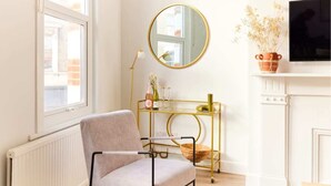 A room with a radiator, a chair, a round mirror, a TV, and a shelf with decorative items. - The Fulham Escape - Alluring 5BDR Flat (London)