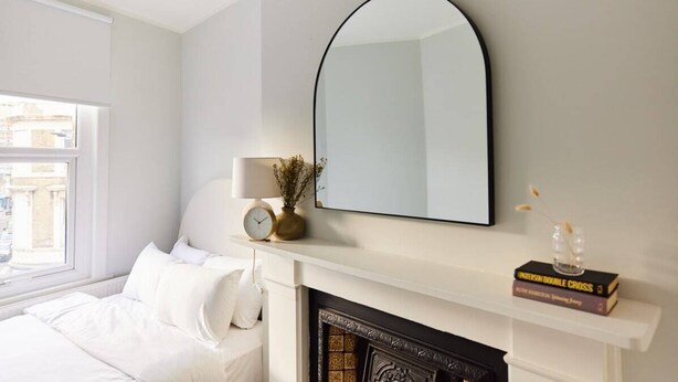 A bedroom with a bed, a white fireplace, a large mirror, and a window. - The Fulham Escape - Alluring 5BDR Flat (London)