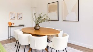 A dining area with a round wooden table surrounded by six white chairs, a vase with plants, and framed artwork on the wall. - The Fulham Escape - Alluring 5BDR Flat (London)