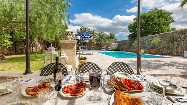 Area pranzo all'aperto con vista sulla piscina, con un tavolo apparecchiato con piatti di cibo, bicchieri e una bottiglia di vino.