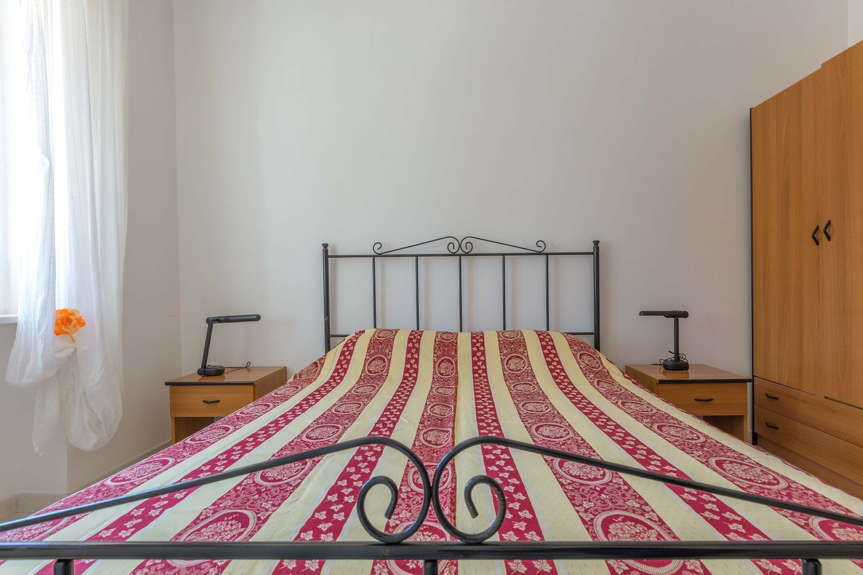 Un dormitorio con estructura de cama de metal, colcha estampada, dos mesitas de noche con lámparas, un armario de madera y una ventana con cortina blanca y una flor naranja.