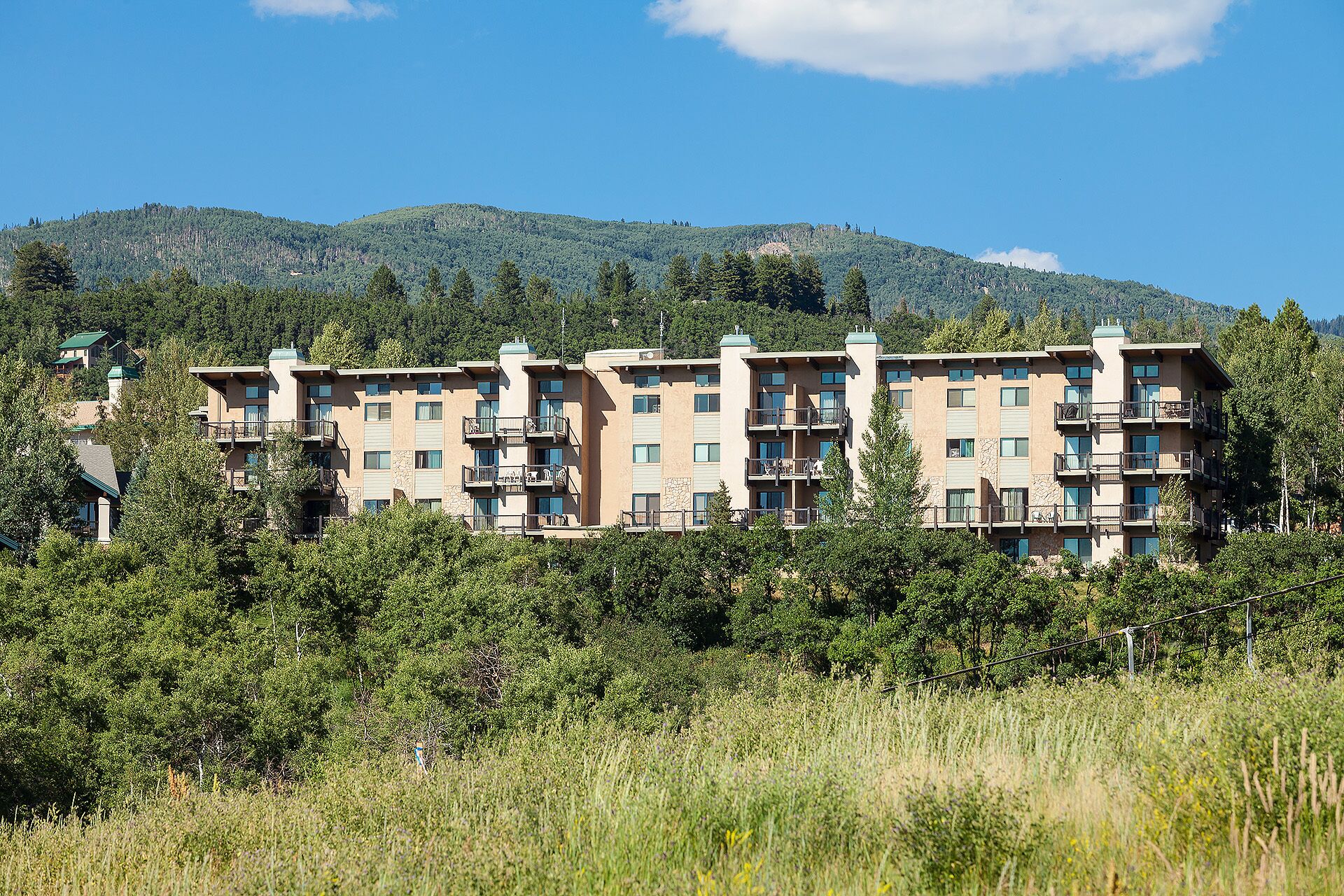 A multi-story hotel with balconies, set against a backdrop of mountains and greenery.