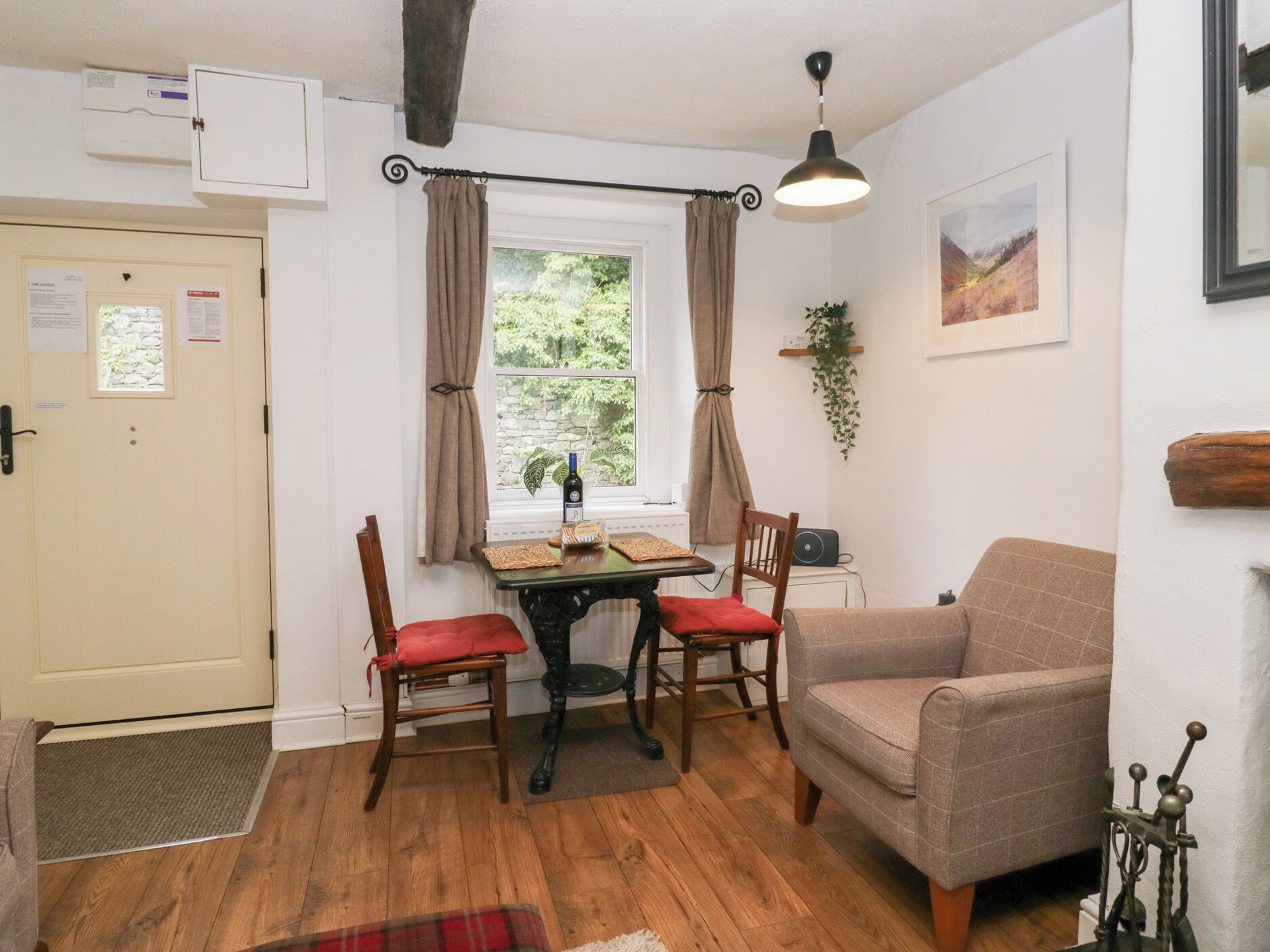 Une pièce chaleureuse avec un plancher en bois, une table à manger avec des chaises, un canapé, un radiateur, une fenêtre avec des rideaux, une plante suspendue et un tableau au mur.