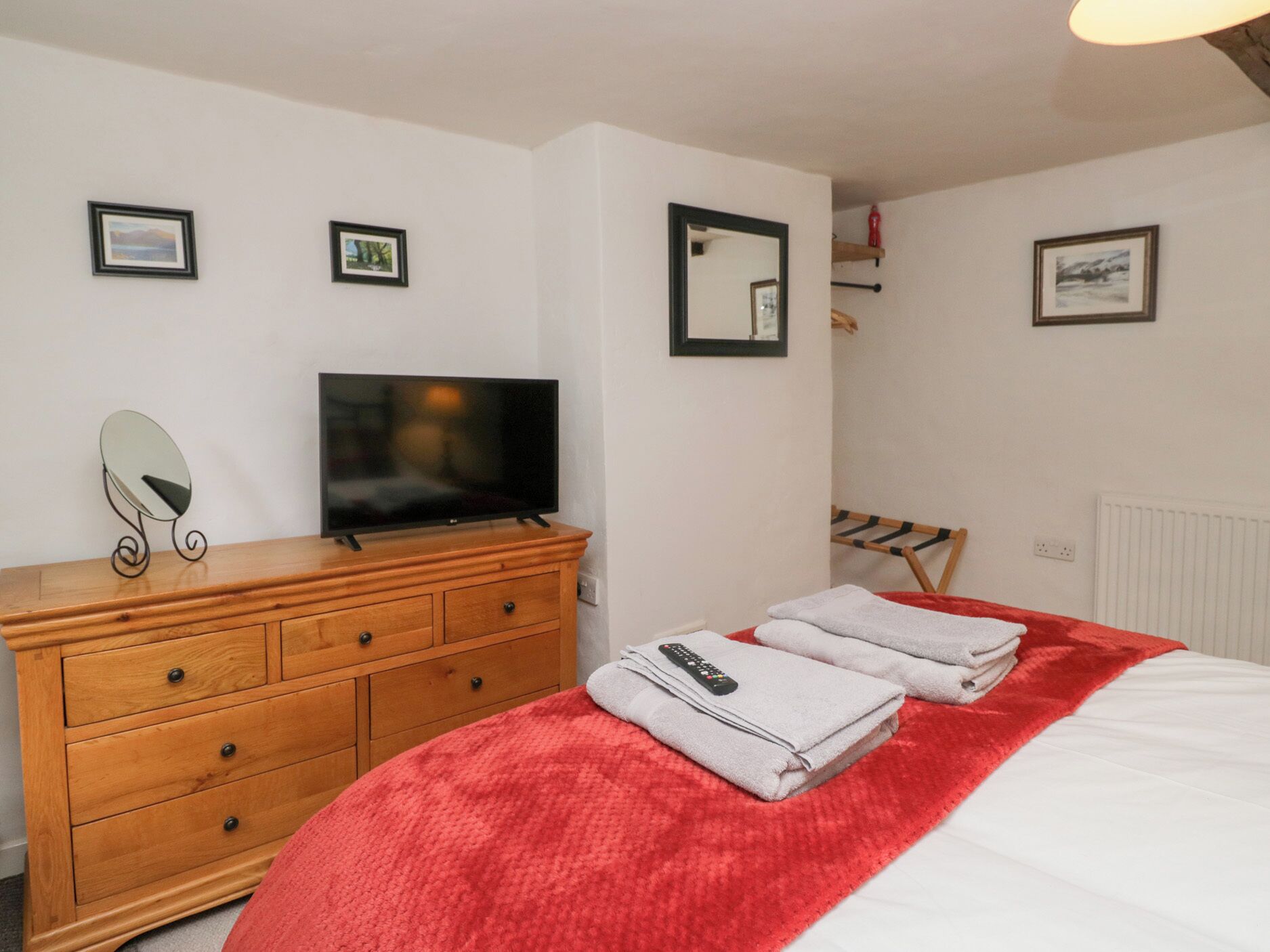 A bedroom with a wooden dresser, a flat-screen TV, a bed with a red blanket, and two framed pictures on the wall.