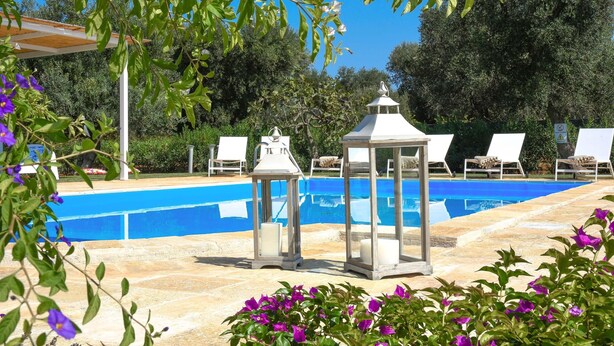 A swimming pool with lounge chairs and lanterns, surrounded by greenery and flowers.