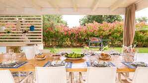 An outdoor dining area with a wooden table set for a meal, surrounded by white wicker chairs.