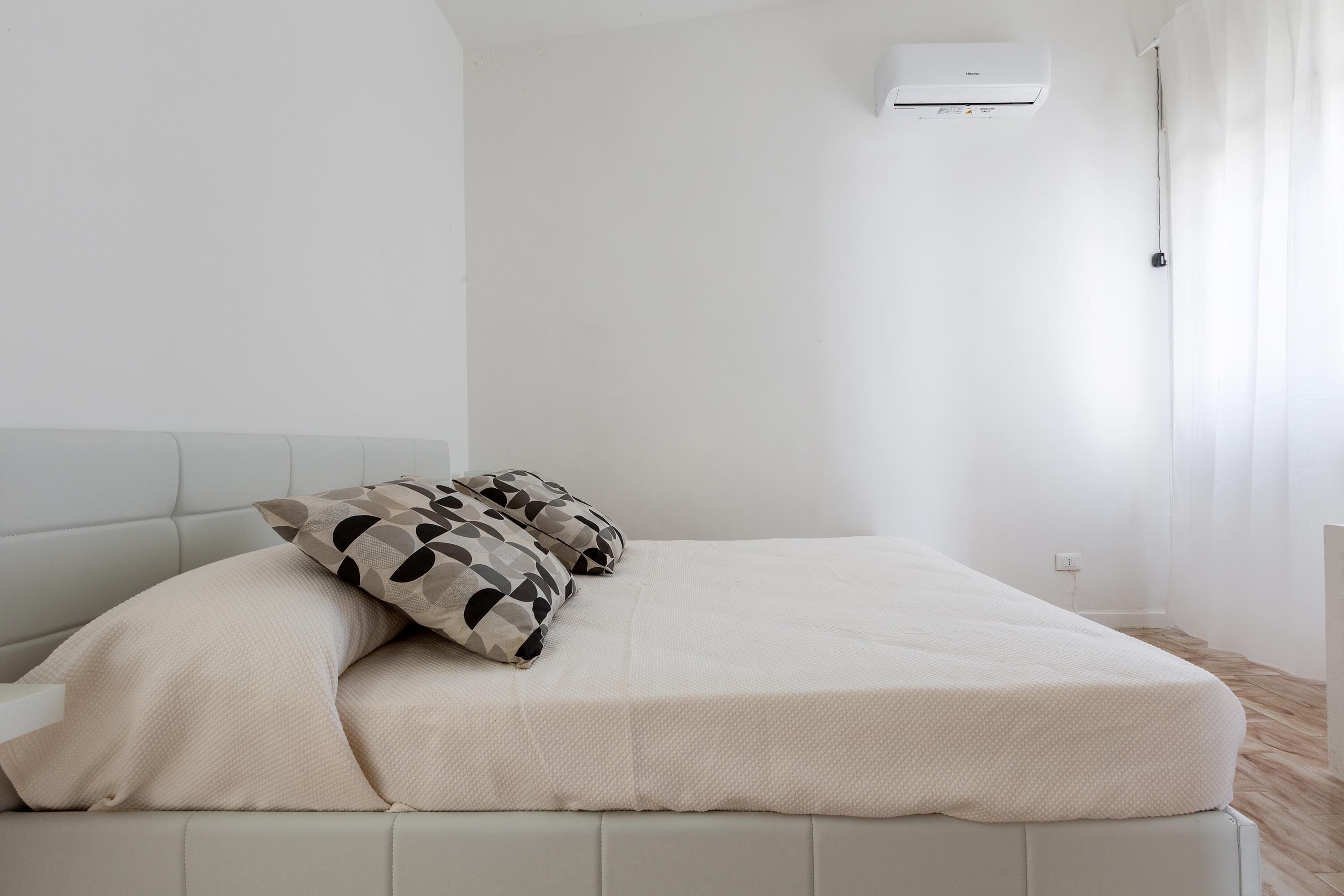 En snyggt bäddad säng med ett beige överkast och mönstrade kuddar, en vit luftkonditioneringsenhet på väggen och ett trägolv.
