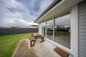 Een terras met een houten tafel en banken, een schuifdeur naar een achtertuin met gazon en omheining.