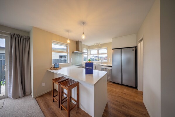 Una cocina moderna con una isla central, taburetes de madera, un refrigerador de acero inoxidable y vistas al exterior a través de grandes ventanales.