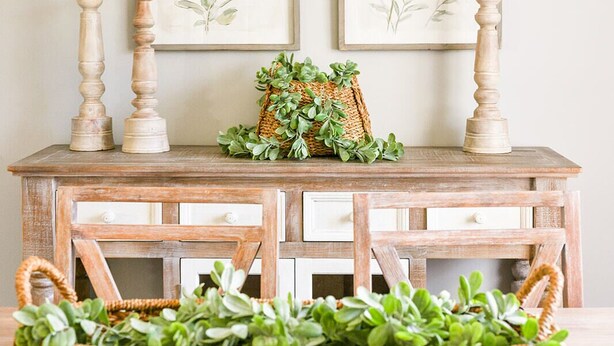 Een houten tafel in rustieke stijl met twee ingelijste botanische prints, een geweven mand met groen en twee kandelaars.
