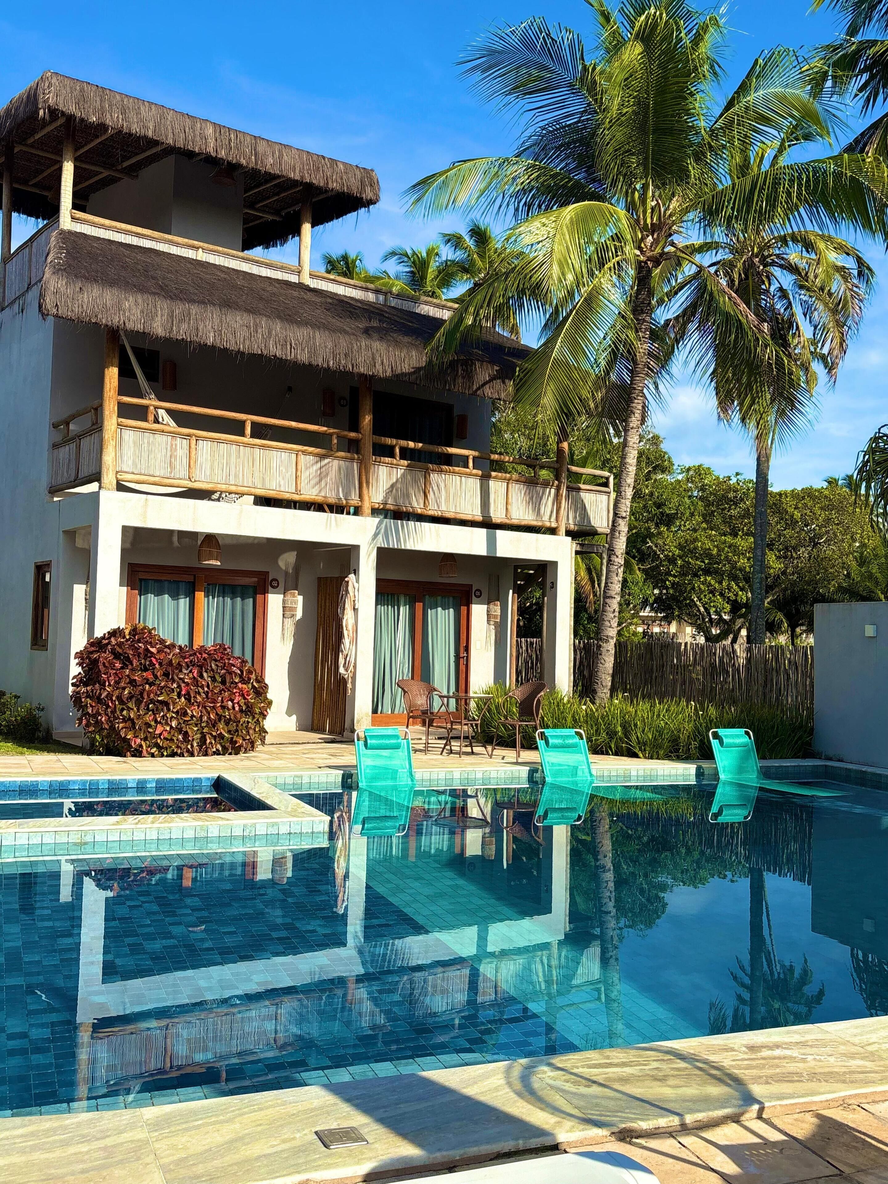 A two-story building with a thatched roof and a pool area with lounge chairs.