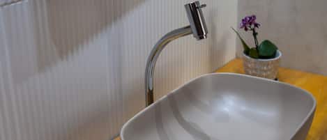 Ein Badezimmer mit einem großen, weißen Waschtisch, einer Holzkommode, einem kleinen Korbwäscheschrank mit Pflegeprodukten und einer Topfpflanzen-Orchidee.