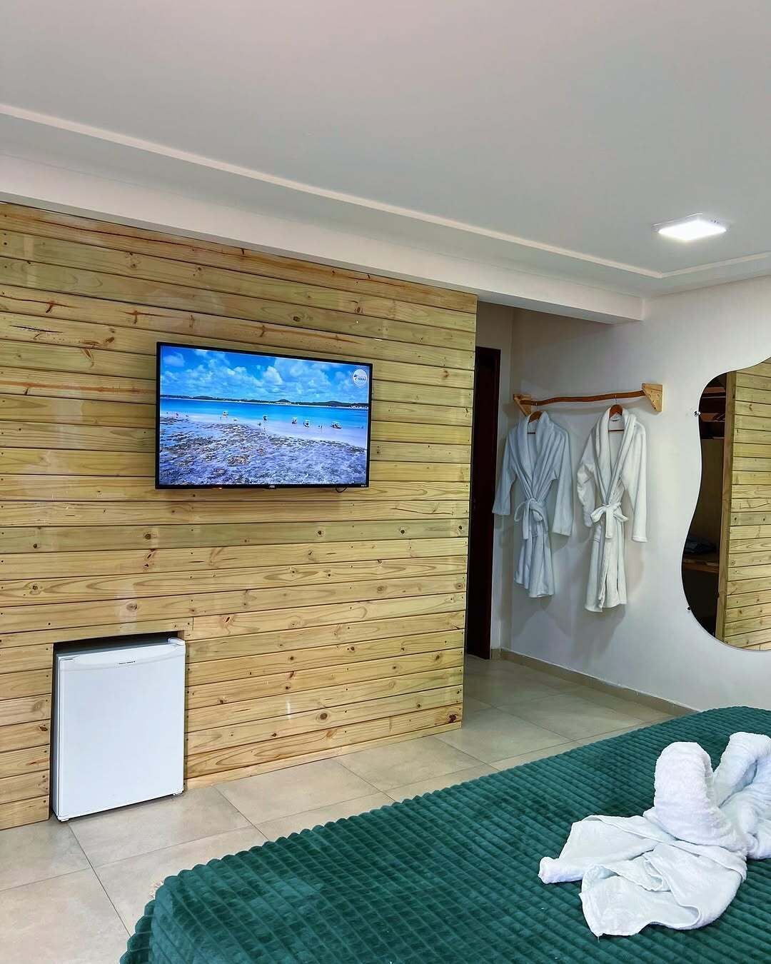 A modern room with a wooden panel wall, a television, a small refrigerator, a green bedspread, and a towel on the floor.