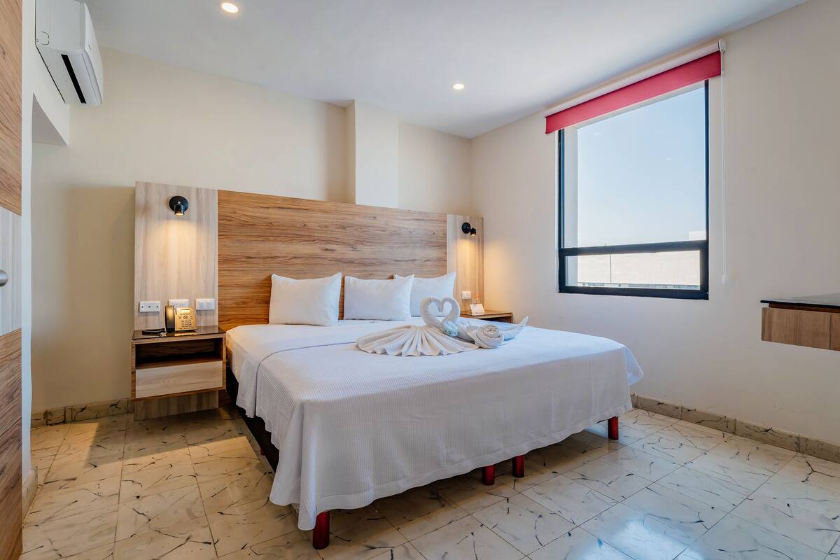 A hotel room with a large bed, bedside tables, a wooden headboard, a window with a red blind, and a tiled floor.