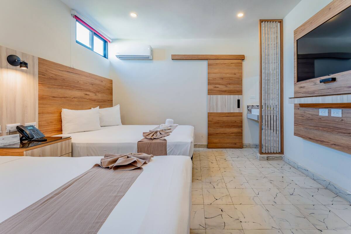 A hotel room with two beds, a wooden headboard, a TV mounted on the wall, a phone on the nightstand, and a window with a red blind.