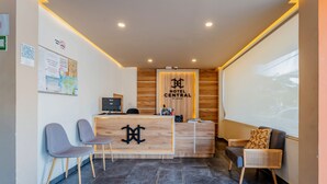 A hotel reception area with a wooden reception desk, two grey chairs, a black computer, and a large window. - Hotel Central Merida by Kavia (Mérida)