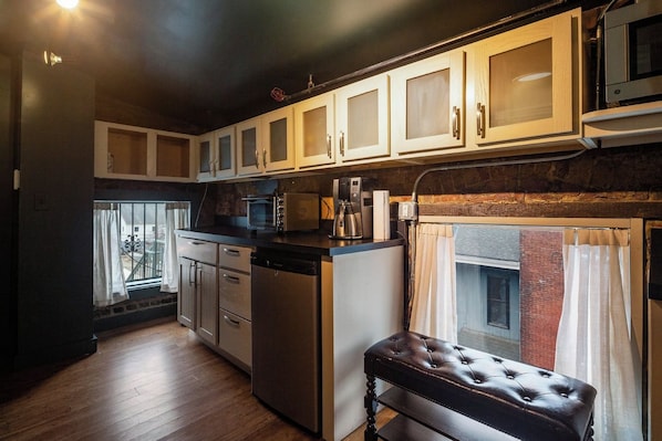 A compact kitchen with white cabinets, a microwave, a coffee maker, and a dishwasher. There is a small bench near the window.