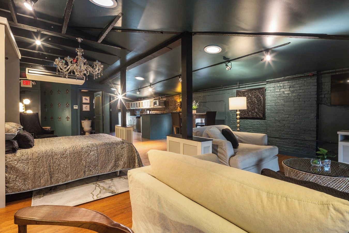 Habitación de hotel moderna con una cama grande, un sofá, una mesita y una cocina visible a través de una puerta abierta.