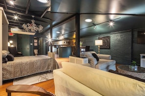 A modern hotel room with a large bed, a sofa, a small table, and a kitchen area visible through an open door.