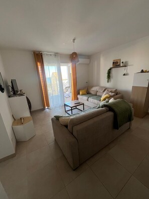 A living room with a sofa, a coffee table, and a TV. There is a sliding door leading to a balcony.
